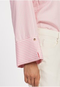 Lichtroze shirt met een geribbelde textuur, voorzien van een brede manchette die is verfraaid met een kleine gouden knoop, gecombineerd met crème-kleurige broek.
