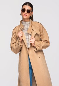 Femme portant un trench-coat marron, un chemisier à col montant à carreaux, un jean bleu, des lunettes de soleil rondes et des boucles d'oreilles en or, tenant le col du manteau.