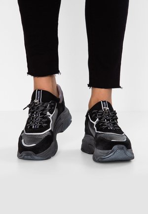 Musta ja harmaa lenkkari, jossa on verkkokankainen ja mokkanahkainen materiaali, paksu pohja, pyöreä muotoilu sekä kuviolliset nauhat, joissa on metallikoristeet.