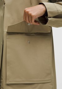 Khaki deštník s hladkou texturou, který má velkou přední kapsu, zapínání na patentky a vyvýšený lem podél okraje.