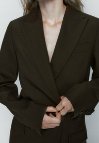 Blazer marron sur mesure avec revers à pointe, manches longues et poignets boutonnés. Comprend une poche poitrine unique et une coupe structurée.