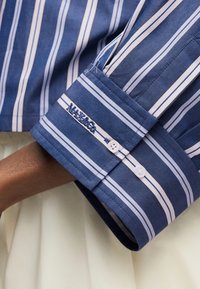 Μανσέτα πουκαμίσου με μπλε και λευκές ρίγες, δύο λευκά κουμπιά και κεντημένο λογότυπο "M&S&Co" σε σκούρο δέρμα, σε ανοιχτό φόντο.