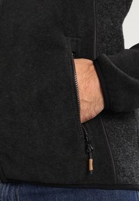Schwarze Fleecjacke mit einem seitlichen Reißverschlusstasche. Verfügt über ein strukturiertes Gewebe, ein glattes Innenfutter und eine Kordel zur Weitenregulierung am Saum.