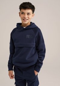 Jūras zilas krūšturis ar teksturētām plecu daļām un ribotu apakšmalu. Priekšpusē ir smalks logotips "BLUE RIDGE DENIM".