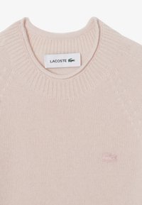 Ljusrosa stickad tröja med ribbad rund halsringning, med en liten broderad Lacoste-logotyp i matchande färg på bröstet.