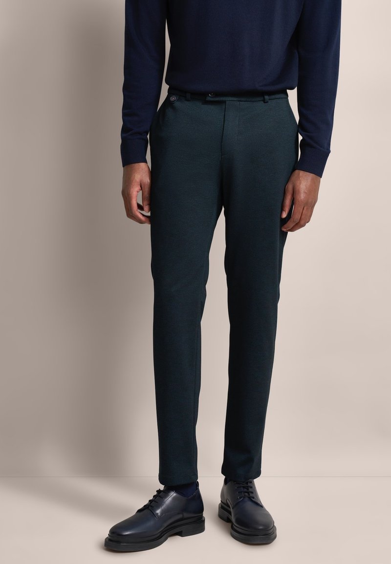 Dunkelgrüne Hose aus strukturiertem Stoff, mit taillierter Passform, Fronttaschen und einem glatten Bund. Kombiniert mit schwarzen Schuhen.
