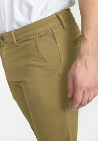 Man met kaki-broek en een wit shirt, hand rustend naast de rechterzak met een klein logo-label zichtbaar.