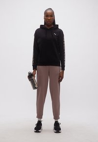 Γυναίκα που φοράει μαύρο φούτερ Puma και βιολετί φόρμα κρατάει διάφανη παγούρι Puma, στέκεται σε λευκό φόντο.