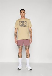 Beežist puuvillast t-särk kamuflaažilogoga, millele on lisatud mauve värvi lühikesed püksid ja valged tossud. Komplekti iseloomustab lõdvestunud lõige ja nähtavad tätoveeringud.