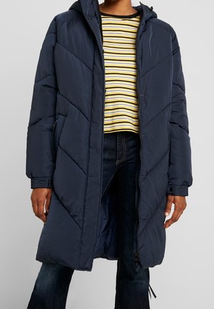 Navy blue puffer coat with a hood, featuring chevron stitching, zippered front, and side pockets, worn over a yellow and black striped shirt.