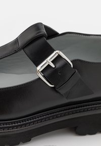 Chaussure en cuir noir avec une bride à boucle argentée et une semelle à bande de roulement épaisse, montrant un gros plan sur la fermeture et les coutures.