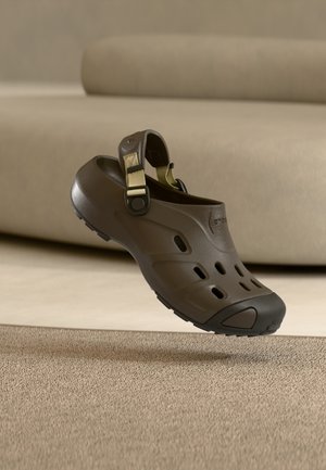 Brauner Slip-On-Clog mit verstellbarem Riemen, Belüftungslöchern und schwarzer Zehenkappe, schwebt über strukturiertem beigem Teppich und Sofahintergrund.