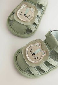 Gröna lädersandaler med en rund, beige björnapplikation på framsidan. Funktioner inkluderar utskärningar och en mjuk textur, idealisk för komfort.