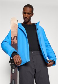 Casaco de esqui azul com fechos e punhos ajustáveis, combinado com calças pretas. O modelo segura esquis castanhos com fixações e detalhes da marca visíveis.