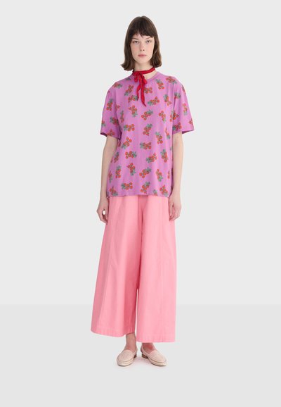 Donna che indossa una camicia floreale rosa, pantaloni rosa larghi, una sciarpa rossa al collo e scarpe rosa chiaro, in piedi davanti a uno sfondo semplice.