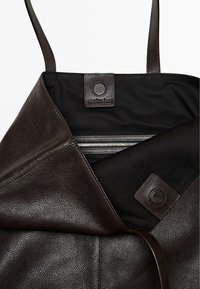 Bruine leren tas met textuur buitenkant, zwarte stofvoering, twee leren handvatten en een ritsvak aan de binnenkant. Kenmerkt een logo-label.