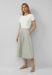 Witte top met korte mouwen, gecombineerd met een lichtgroene, geplooide midi-rok met zakken. Model draagt beige sandalen met decoratieve banden.