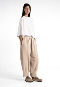 T-shirt blanc surdimensionné, pantalon beige avec ceinture élastique, et chaussures brunes à enfiler. Le modèle se tient avec les mains dans les poches sur un fond blanc.