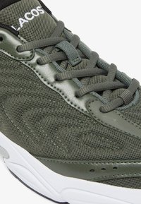 Sneaker verde oliva con materiali in rete e sintetici, suola intermedia texturizzata, lacci abbinati e logo sul collo. Contrasto con suola bianca.