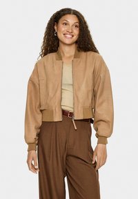 Frau mit lockigem Haar, die eine beigefarbene Jacke über einem beigen Shirt und hochtaillierten braunen Hosen trägt, lächelt und steht vor einem schlichten Hintergrund.