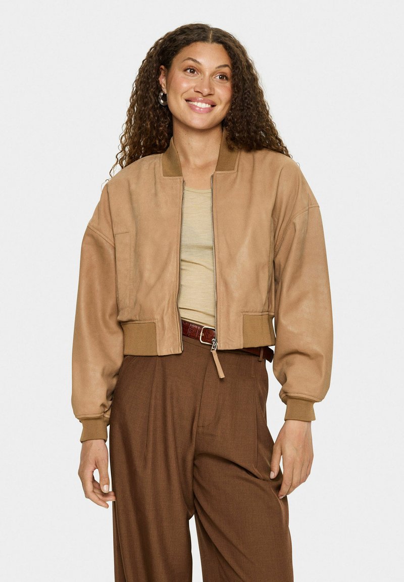 Frau mit lockigem Haar, die eine beigefarbene Jacke über einem beigen Shirt und hochtaillierten braunen Hosen trägt, lächelt und steht vor einem schlichten Hintergrund.