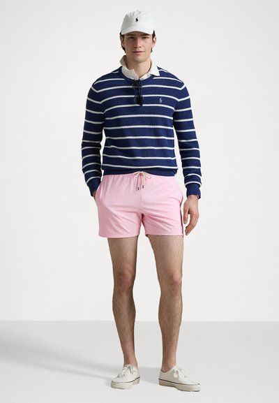Pullover în dungi albastre și albe, pantaloni de baie roz cu șnur, șapcă albă și pantofi casual. Ochelari de soare așezați pe pulover.