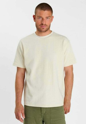 Man met kort haar die een gestructureerd gebroken wit shirt met korte mouwen en olijfgroene broek draagt, staand tegen een effen witte achtergrond.