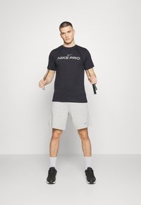 Nike Pro lühikeste varrukatega must T-särk koos hallide kergete šortsidega. Mudel kannab musti kvaliteetseid jooksujalatseid ja valgeid sokke, hoides käes hüppenöör.