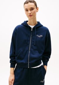 Hoodie zippé marine en tissu doux, avec une poche kangourou, des poignets côtelés et un petit logo sur la poitrine. Associé à un bas coordonné.