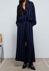 Schwarz-blau gestreifter Overall mit floralem Muster, langen Ärmeln, lockerer Passform und Kordelzug in der Taille. Kombiniert mit schwarzen High Heels.