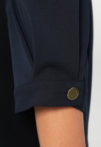 Tumši zila krekla piedurkne ar apvītu apmali un zeltīta pogas akcentu uz aproces. Audums izskatās gluds un nedaudz fakturēts.