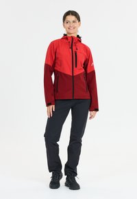 Red and burgundy waterproof jacket with a hood, black zipper pockets, elastic cuffs, paired with black pants and sturdy black boots.