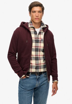 Felpa bordeaux con zip e cappuccio regolabile indossata sopra una camicia flanella a quadri nei toni beige, blu e rosso. Jeans blu completano l'outfit.
