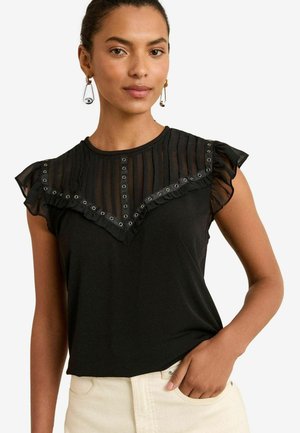 Woman with medium skin tone, wearing a black top with ruffled sleeve details and decorative buttons, styled against a white background.