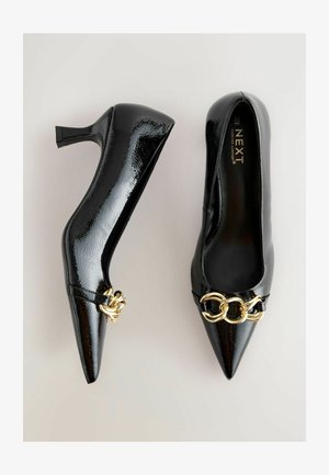 Schwarze Lackleder High Heels mit spitzen Zehen und einem dekorativen goldenen Kettendetail an der Vorderseite, vor einem hellen Hintergrund.