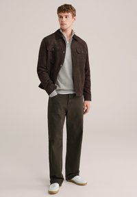 Geacă maro din piele întoarsă cu două buzunare la piept, pulover gri tricotat, pantaloni verde închis și adidași albi cu talpă bej. Design simplu și structurat.