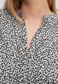 Zwart-witte blouse met een v-hals en een korte sluiting. Heeft een bloemendessin met kleine, verspreide vormen. Gladde stofstructuur.