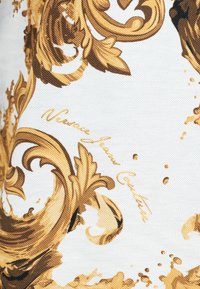 Σχέδιο με χρυσά στυλιζαρισμένα scrolls και φύλλα σε μπαρόκ στυλ πάνω σε λευκό ύφασμα με καλλιγραφική γραφή "Versace Jean Couture".