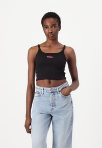Top corto nero con spalline sottili, con un piccolo logo rosa al centro. Abbinato a jeans azzurri a vita alta.