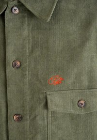 Chaqueta de pana en color verde oliva con botones marrones, un bolsillo en el pecho y una pequeña "P" bordada en rojo con diseño floral.