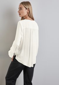 Blouse blanche à manches longues en tissu léger, avec un pli au dos, un col rond et des poignets boutonnés, associée à un jean noir.