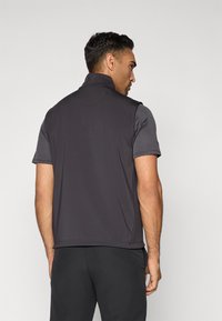 Man gezien vanuit de rug met een donker mouwloos vest over een T-shirt met korte mouwen en zwarte broek tegen een effen lichte achtergrond.
