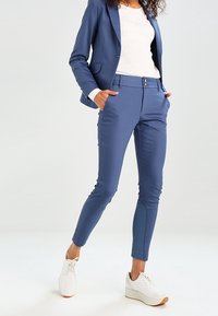 Modrý řezaný blejzer a odpovídající slim-fit kalhoty z hladkého materiálu. Kalhoty mají dvě přední kapsy a zapínání na knoflík. Kombinováno s bílými teniskami.