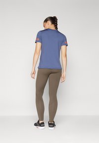 Kék, narancssárga márkajelzéssel ellátott sportpóló, barna leggings-szel és fekete sportcipővel párosítva, hátulról nézve.