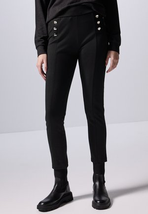 Zwarte slim fit broeken met zijknopen, gladde stof, enkel-lange ontwerp, gecombineerd met zwarte enkellaarsjes en een donkere trui.