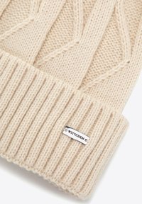Berretto a maglia color beige chiaro con motivo lavorato a trama e bordo a costine. Presenta un'etichetta con logo in argento cucita sul bordo.