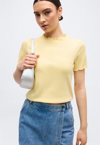 Νεαρή γυναίκα που φοράει κίτρινο τοπ με βολάν στις άκρες, φούστα από τζιν και ασημένια τσάντα ώμου, πόζαρε μπροστά σε απλό λευκό φόντο.