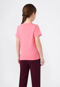 Κορίτσι με καστανά πλεγμένα μαλλιά, φορώντας ροζ μπλούζα κοντού μανικιού και σκούρους μωβ παντελόνες, στέκεται γυρισμένο προς τα πίσω μπροστά σε απλή φόντο.