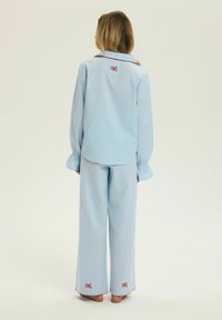 Osoba stojąca boso, ubrana w jasnoniebieską piżamę z czerwonymi haftowanymi kokardami na plecach i nogawkach, neutralne tło.