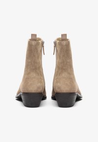 Botas de tobillo de ante beige con un tacón corto en bloque, que cuentan con un cierre de cremallera en la parte trasera y un tirador decorativo. Textura suave, diseño clásico.